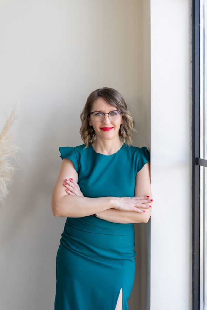 Júlia virtuális asszisztens zöld ruhában, karba tett kézzel az ablak mellett