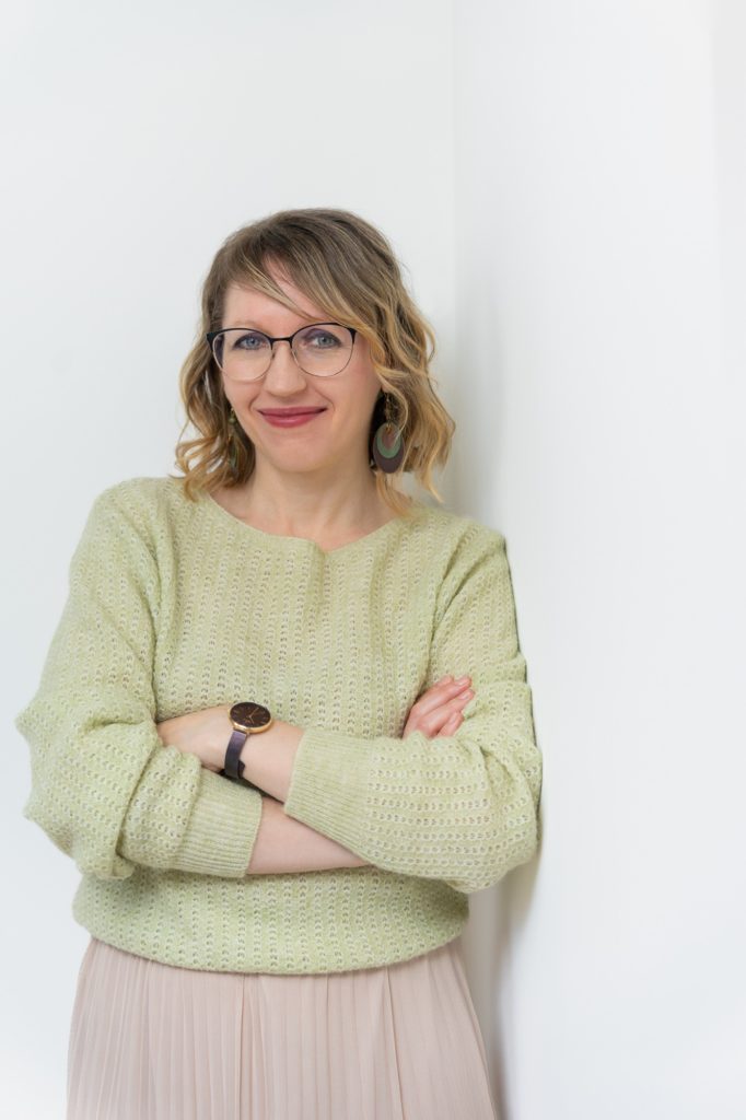 Júlia virtuális asszisztens zöld pulóverben, karba tett kézzel a falnak dől