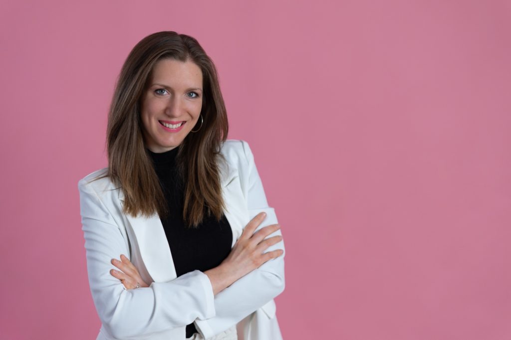 Regi webdesigner fehér blézerben, karba tett kézzel egy rózsaszín háttér előtt