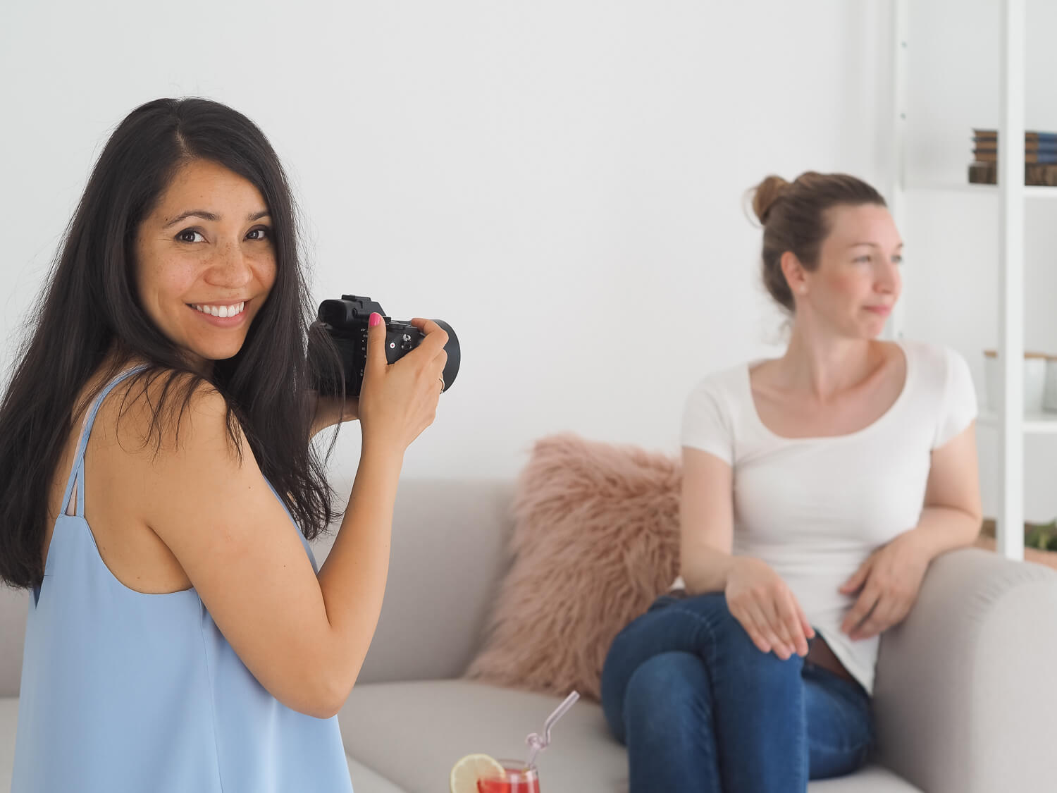 Fotós nő fényképezőgéppel mosolyog, mögötte egy nő ül a kanapén – brand fotográfus fotózása