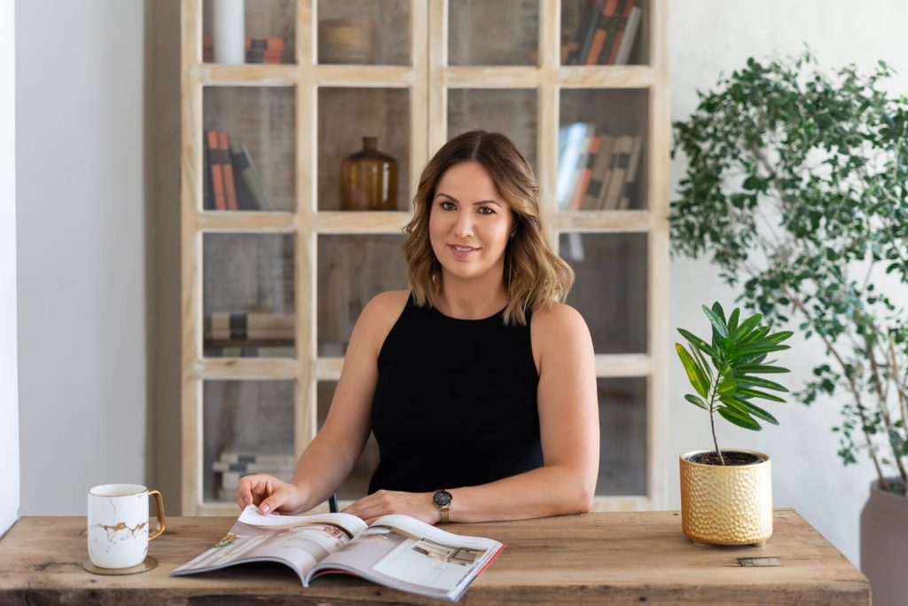 Vállalkozónő fekete felsőben magazint lapoz egy asztalnál, mögötte könyvespolc
