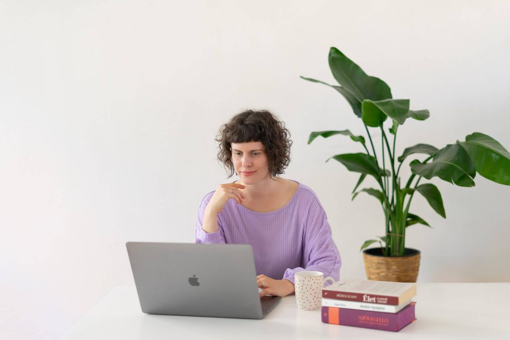 Rövid hajú vállalkozónő lila pulóverben laptopon dolgozik, mellette nagy szobanövény